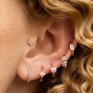 May include: A close-up of an ear adorned with multiple pieces of jewelry. The ear features a small flower stud, a small hoop with clear stones, a small hoop with a four-petal flower, a hoop with a butterfly design, a hoop with a flower design, and a rectangular earring.