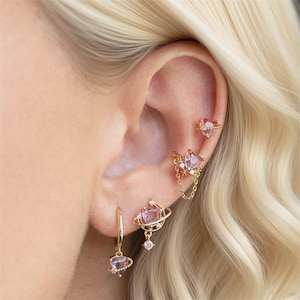 Può includere: Una collezione di orecchini dorati con motivi a cuore, pianeta e stella, adornati con pietre preziose rosa e trasparenti. Gli orecchini includono cerchi, borchie e un ear cuff a catena, che presentano un tema celeste.