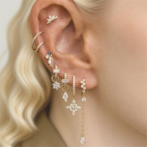 Puede incluir: Una variedad de pendientes dorados con diversos diseños, incluyendo tachuelas, aros y estilos colgantes. Los pendientes presentan piedras preciosas claras y azules, y formas florales y geométricas. Las joyas se muestran en una oreja.