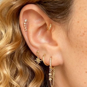 Könnte beinhalten: Nahaufnahme eines Ohrs, das mit mehreren goldenen Ohrringen geschmückt ist. Ein Helix-Piercing zeigt einen gebogenen Stab mit kleinen, funkelnden Steinen. Ein Creolen-Ohrring und ein baumelnder Stern-Ohrring sind ebenfalls sichtbar, zusammen mit einem Ketten-Ohrring mit drei baumelnden Edelsteinen.