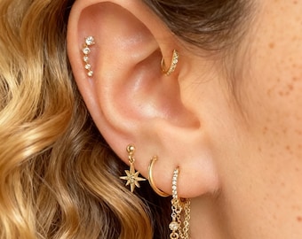 Könnte beinhalten: Nahaufnahme eines Ohrs, das mit mehreren goldenen Ohrringen geschmückt ist. Ein Helix-Piercing zeigt einen gebogenen Stab mit kleinen, funkelnden Steinen. Ein Creolen-Ohrring und ein baumelnder Stern-Ohrring sind ebenfalls sichtbar, zusammen mit einem Ketten-Ohrring mit drei baumelnden Edelsteinen.