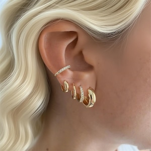 Peut inclure: Gros plan sur une oreille ornée de plusieurs boucles d'oreilles dorées. L'ensemble comprend de petits cerceaux, un cerceau plus grand, un cerceau délicat avec de petites pierres et une boucle d'oreille à manchette avec de petites pierres.