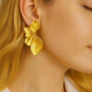 Peut inclure: Boucles d'oreilles florales dorées. Les boucles d'oreilles présentent un design texturé en trois dimensions ressemblant à des pétales de fleurs stylisés. Les boucles d'oreilles sont portées à l'oreille d'une personne aux cheveux blonds.