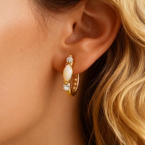 May include: Gold hoop earrings with a central oval-shaped white stone and two smaller clear stones. The earrings are set in a gold metal and are worn on an ear. The background is blurred.