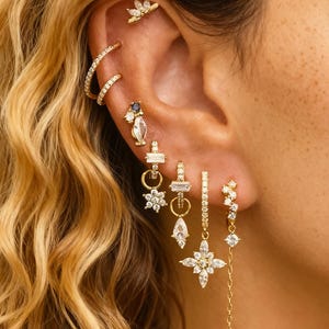May include: A collection of gold-toned earrings adorns an ear. The earrings include hoops, studs, and dangling styles, all embellished with clear and blue gemstones. A small, delicate floral stud is placed at the top of the ear.