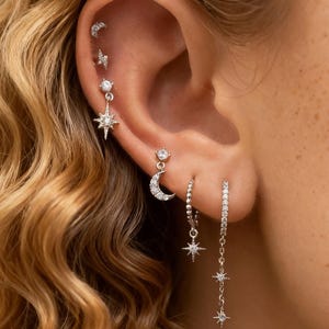 Könnte beinhalten: Ein Ohr mit mehreren silbernen Ohrringen. Die Ohrringe umfassen einen Blitzbolzen-Stecker, einen sternförmigen Anhänger, einen Halbmond-Stecker, einen Reifen mit einem hängenden Stern und eine lange Kette mit drei Sternen. Der Schmuck ist mit klaren Edelsteinen besetzt.