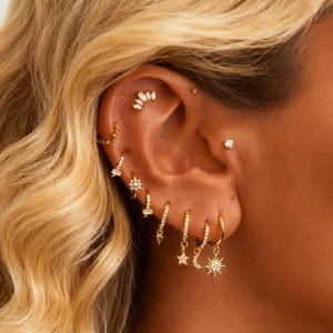 Op de afbeelding: Close-up van een oor versierd met meerdere gouden oorbellen. Het oor heeft een mix van studs, hoepels en bungelende oorbellen met verschillende ontwerpen, waaronder sterren, manen en kristallen. De oorbellen zijn geplaatst tegen een achtergrond van blond haar.