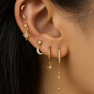 Op de afbeelding: Een verzameling gouden oorbellen met maan-, ster- en sterrenmotieven. De oorbellen worden op een oor gedragen en tonen verschillende stijlen, waaronder studs, creolen en bungelende oorbellen. De sieraden zijn versierd met kleine, sprankelende details.