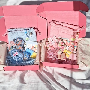 May include: Two pink gift boxes filled with assorted items, including pouches, notebooks, and hair accessories. The boxes are lined with patterned paper and filled with blue and pink decorative filler. The items are in shades of pink, blue, and yellow.