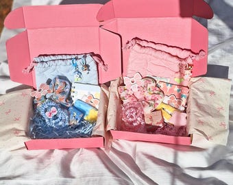 May include: Two pink gift boxes filled with assorted items, including pouches, notebooks, and hair accessories. The boxes are lined with patterned paper and filled with blue and pink decorative filler. The items are in shades of pink, blue, and yellow.