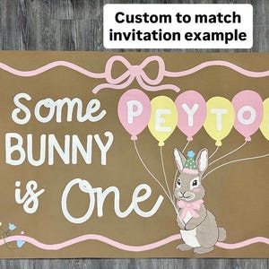 May include: A rectangular brown sign featuring the white text Some BUNNY is One. A gray bunny wearing a party hat and pink bow holds five balloons in pink and yellow, each letter of PEYTON printed on them. A pink border and floral accents.