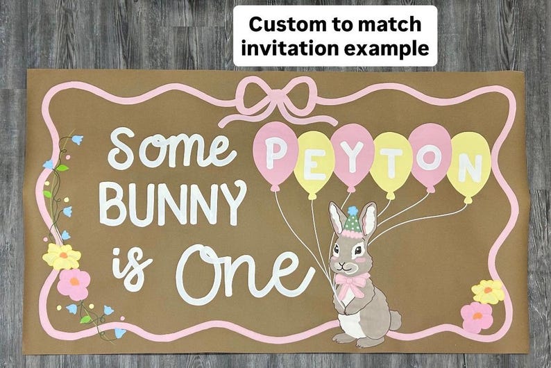 May include: A rectangular brown sign featuring the white text Some BUNNY is One. A gray bunny wearing a party hat and pink bow holds five balloons in pink and yellow, each letter of PEYTON printed on them. A pink border and floral accents.