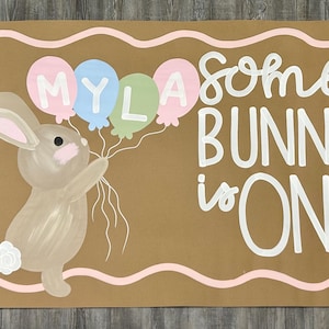 May include: A rectangular tan sign with a wavy pink border featuring a brown bunny holding four balloons labeled MYLA. The text reads Some BUNNY is ONE in white script and block letters. A pink rose and blue flowers are on the right side.