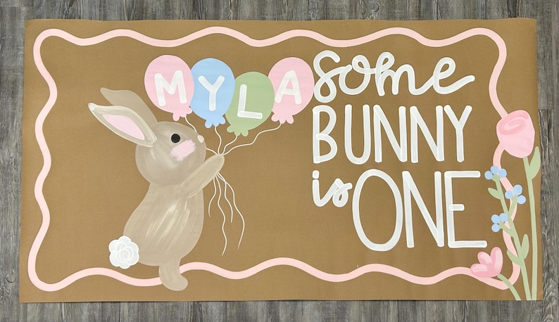 May include: A rectangular tan sign with a wavy pink border featuring a brown bunny holding four balloons labeled MYLA. The text reads Some BUNNY is ONE in white script and block letters. A pink rose and blue flowers are on the right side.