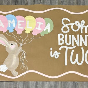 May include: A rectangular brown paper banner featuring a hand-painted illustration of a light brown bunny holding a cluster of pastel-colored balloons with the name AMELIA written on them. The text Some BUNNY is TWO is written in white.