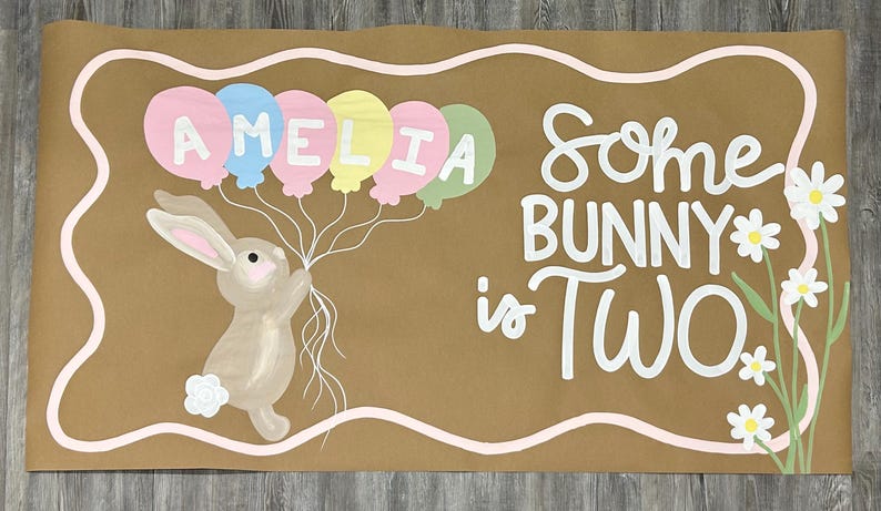 May include: A rectangular brown paper banner featuring a hand-painted illustration of a light brown bunny holding a cluster of pastel-colored balloons with the name AMELIA written on them. The text Some BUNNY is TWO is written in white.
