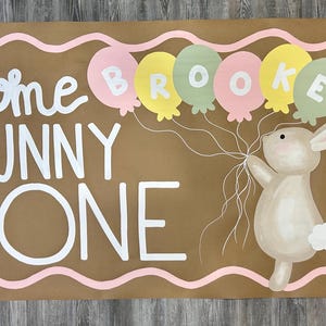 May include: A rectangular brown paper banner featuring the white text Some BUNNY is ONE. A tan bunny holds a cluster of pastel pink, yellow, and sage green balloons with the name BROOKE written on them. A pink wavy border and floral accents frame the design.