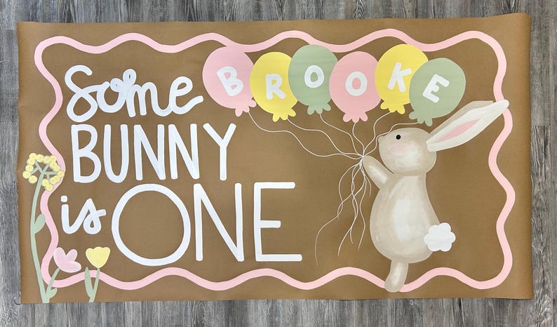 May include: A rectangular brown paper banner featuring the white text Some BUNNY is ONE. A tan bunny holds a cluster of pastel pink, yellow, and sage green balloons with the name BROOKE written on them. A pink wavy border and floral accents frame the design.