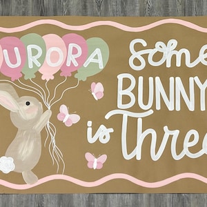 May include: A rectangular brown sign featuring a painted bunny holding a cluster of balloons with the name AURORA written on them. The text reads Some BUNNY is Three, accompanied by white daisies, pink butterflies, and a wavy pink border.