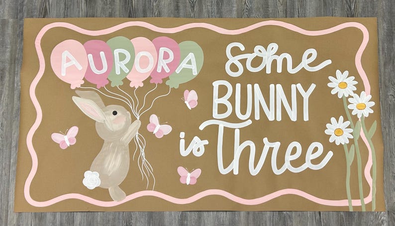 May include: A rectangular brown sign featuring a painted bunny holding a cluster of balloons with the name AURORA written on them. The text reads Some BUNNY is Three, accompanied by white daisies, pink butterflies, and a wavy pink border.
