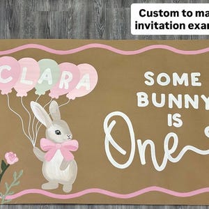 May include: A rectangular brown sign with a pink wavy border featuring a white rabbit wearing a pink bow, holding a cluster of pink and green balloons with the name CLARA written on them, and the white text SOME BUNNY IS One with a bow graphic.