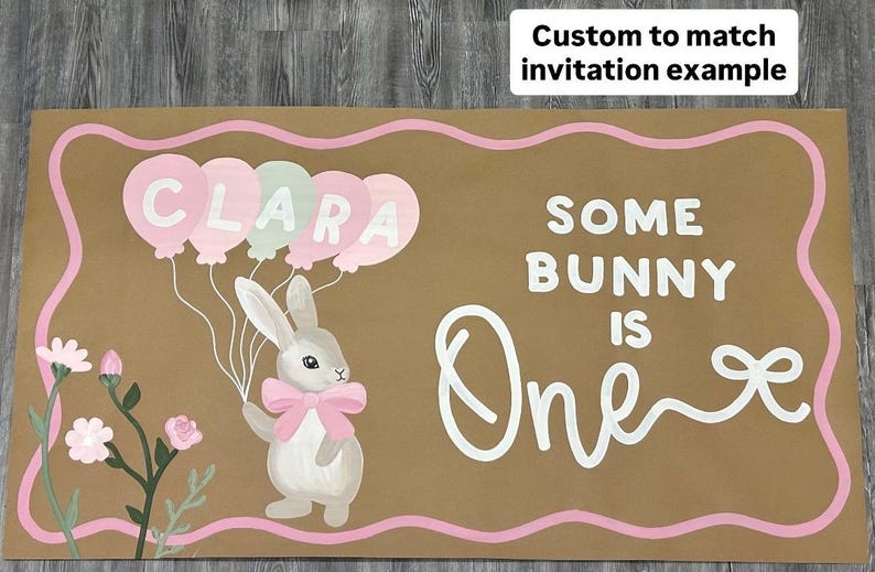 May include: A rectangular brown sign with a pink wavy border featuring a white rabbit wearing a pink bow, holding a cluster of pink and green balloons with the name CLARA written on them, and the white text SOME BUNNY IS One with a bow graphic.