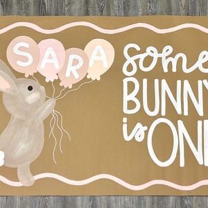May include: A rectangular tan sign featuring a painted gray bunny holding four pastel balloons with the name SARA written on them. The text reads some BUNNY is ONE in white lettering, accompanied by white daisy flowers and a wavy pink border.