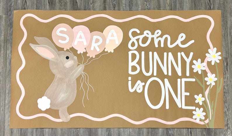 May include: A rectangular tan sign featuring a painted gray bunny holding four pastel balloons with the name SARA written on them. The text reads some BUNNY is ONE in white lettering, accompanied by white daisy flowers and a wavy pink border.