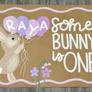 May include: A rectangular brown sign featuring a painted tan bunny holding a cluster of purple and lavender balloons with the name RAYA written on them. The text reads SOME BUNNY IS ONE in white, surrounded by painted green foliage, purple flowers, and butterflies.