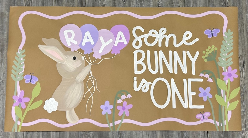 May include: A rectangular brown sign featuring a painted tan bunny holding a cluster of purple and lavender balloons with the name RAYA written on them. The text reads SOME BUNNY IS ONE in white, surrounded by painted green foliage, purple flowers, and butterflies.