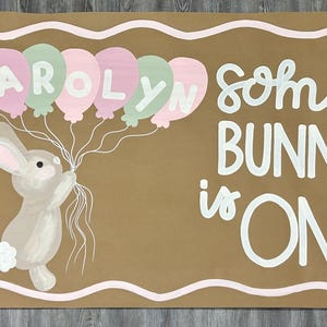 May include: A rectangular brown sign featuring a painted illustration of a light brown bunny holding a cluster of pink and mint green balloons with the name CAROLYN written on them. The text reads SOME BUNNY is ONE, accompanied by a pink flower.