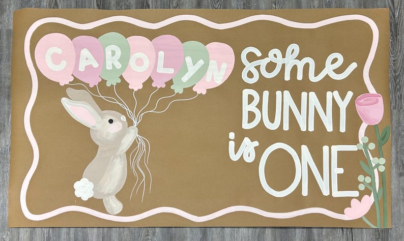 May include: A rectangular brown sign featuring a painted illustration of a light brown bunny holding a cluster of pink and mint green balloons with the name CAROLYN written on them. The text reads SOME BUNNY is ONE, accompanied by a pink flower.