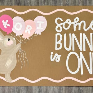 May include: A rectangular tan sign featuring a painted brown bunny holding three balloons with the name KORI written on them. The text reads some BUNNY is ONE in white lettering. A pink wavy border frames the sign, and pink flowers appear on the right.