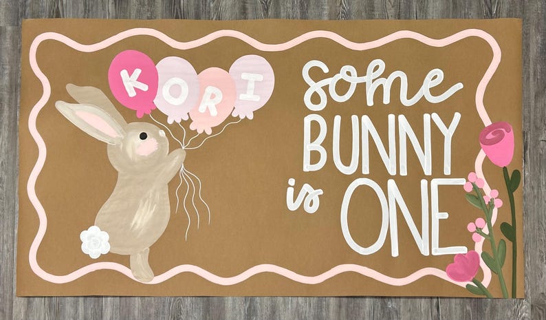 May include: A rectangular tan sign featuring a painted brown bunny holding three balloons with the name KORI written on them. The text reads some BUNNY is ONE in white lettering. A pink wavy border frames the sign, and pink flowers appear on the right.
