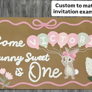 May include: A rectangular brown sign featuring the white text Some Bunny Sweet is One, a pink bow border, a bunny with a pink bow, and balloons spelling VICTORIA in alternating pink and light green. The sign includes floral illustrations.