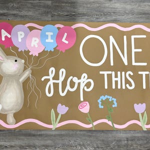 May include: A rectangular brown sign with a pink wavy border featuring a painted gray rabbit holding four balloons labeled APRIL. The text reads ONE HOP THIS TIME in white lettering, accompanied by painted flowers in pink, purple, and blue hues.
