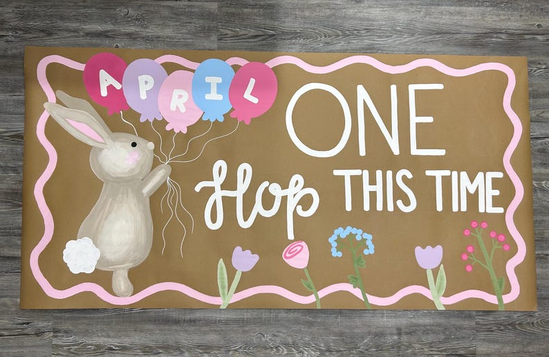 May include: A rectangular brown sign with a pink wavy border featuring a painted gray rabbit holding four balloons labeled APRIL. The text reads ONE HOP THIS TIME in white lettering, accompanied by painted flowers in pink, purple, and blue hues.