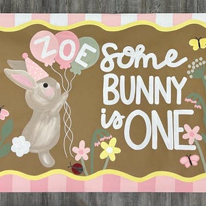 May include: A rectangular sign with a pink and white gingham border featuring a brown center panel. It displays a painted bunny wearing a party hat, holding balloons labeled ZOE, and the text SOME BUNNY IS ONE, surrounded by flowers and butterflies.