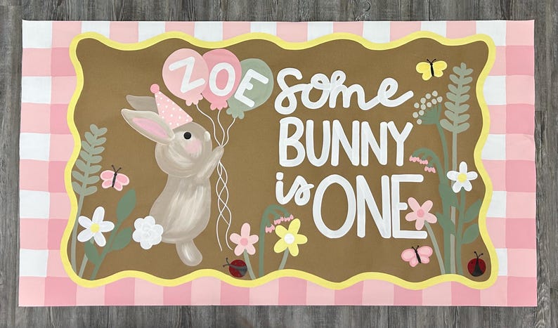 May include: A rectangular sign with a pink and white gingham border featuring a brown center panel. It displays a painted bunny wearing a party hat, holding balloons labeled ZOE, and the text SOME BUNNY IS ONE, surrounded by flowers and butterflies.