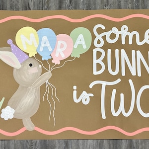 May include: A rectangular brown sign featuring a painted tan bunny wearing a purple party hat and holding three pastel balloons with the name MARA written on them. The text reads some BUNNY is TWO in white, surrounded by painted flowers.
