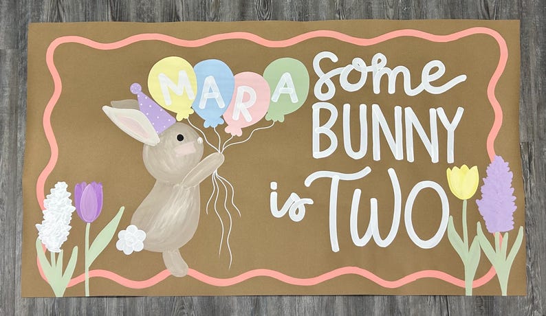 May include: A rectangular brown sign featuring a painted tan bunny wearing a purple party hat and holding three pastel balloons with the name MARA written on them. The text reads some BUNNY is TWO in white, surrounded by painted flowers.