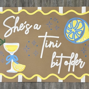 May include: Brown rectangular sign with the text "She's a Tini bit ofter" in white script. The sign features a yellow and blue lemon slice, a cocktail glass with a blue bow, and starbursts.