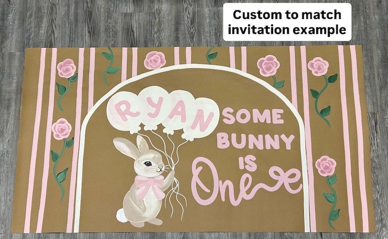 May include: A rectangular sign with a tan background featuring vertical pink stripes and pink roses on vines. A white arch frames a bunny holding balloons with the name RYAN and the text SOME BUNNY IS ONE. A text box reads Custom to match invitation example.