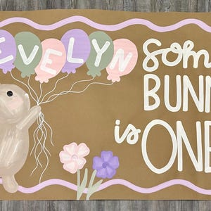 May include: A rectangular brown banner featuring a painted illustration of a tan bunny holding a cluster of pastel balloons. The balloons spell the name EVELYN. The text SOME BUNNY is ONE is written in white, surrounded by flowers and a wavy border.