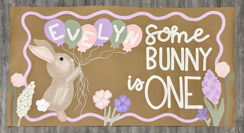 May include: A rectangular brown banner featuring a painted illustration of a tan bunny holding a cluster of pastel balloons. The balloons spell the name EVELYN. The text SOME BUNNY is ONE is written in white, surrounded by flowers and a wavy border.