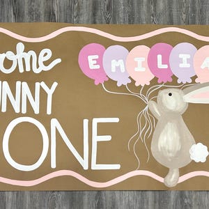 May include: A rectangular brown sign with a wavy pink border featuring the white text Some BUNNY is ONE. A gray bunny holds a cluster of pink and purple balloons with the name EMILIA written in white letters. Pink flowers on green stems are on the left.