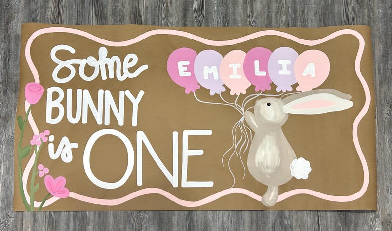 May include: A rectangular brown sign with a wavy pink border featuring the white text Some BUNNY is ONE. A gray bunny holds a cluster of pink and purple balloons with the name EMILIA written in white letters. Pink flowers on green stems are on the left.