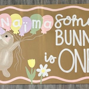 May include: A rectangular brown sign with a wavy pink border featuring a painted brown bunny wearing a purple polka-dot party hat. The bunny holds five pastel balloons with the name Naima written on them. The text reads some BUNNY is ONE.