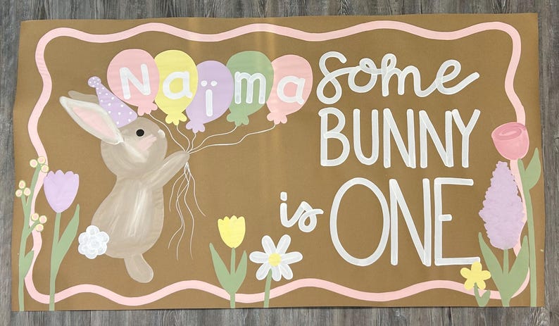 May include: A rectangular brown sign with a wavy pink border featuring a painted brown bunny wearing a purple polka-dot party hat. The bunny holds five pastel balloons with the name Naima written on them. The text reads some BUNNY is ONE.