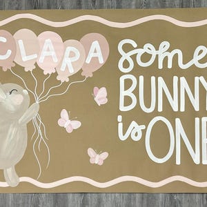 May include: A rectangular tan sign featuring a painted illustration of a light brown bunny holding five pink balloons with the name CLARA written on them. The text reads SOME BUNNY is ONE, accompanied by three small pink butterflies and white daisies.